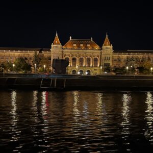 Opplyst historisk bygning langs Donau i Budapest om natten, med gylne lys som speiler seg i elven.